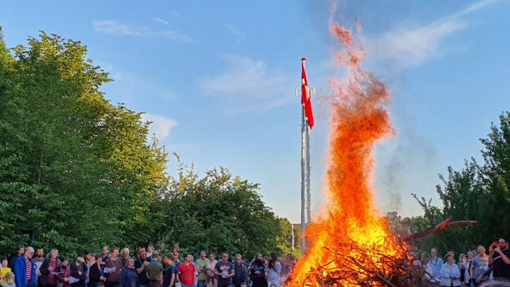 Et stort bål raser, en flok ser med, Dannebrog flyver fra en flagstang i baggrunden