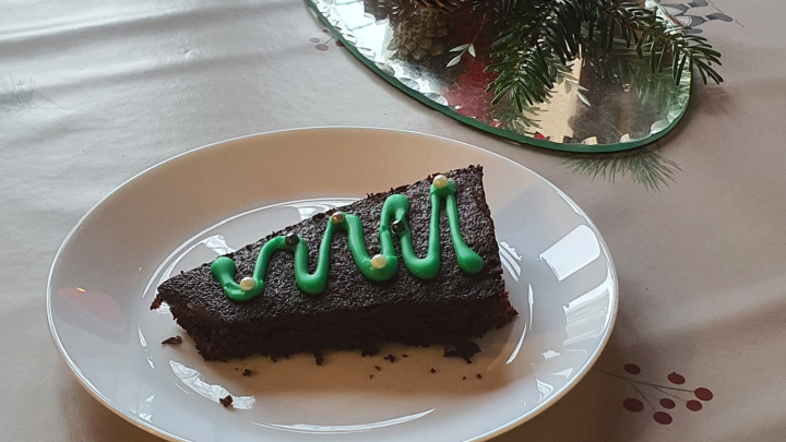 Et stykke chokoladekage formet som et juletræ ligger på en hvid tallerken. Kagen er dekoreret med grøn glasur i form af et juletræ. Ved siden af kagen står et tændt rødt stearinlys og nogle fyrrekogler på et sølvfad. Bordet er dækket med et hvidt dug med et julemønster.