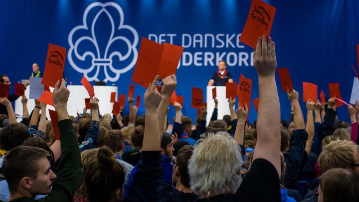 En flok mennesker sætter sig i et auditorium, de fleste af dem holder et rødt stemmekort i luften med den ene arm. På scenen står der nogen ved et podie, bag dem er en blå væg med spejderlogoet og man kan se, at Det Danske Spejderkorps er skrevet, men mange af stemmekortene spærrer for den fulde tekst