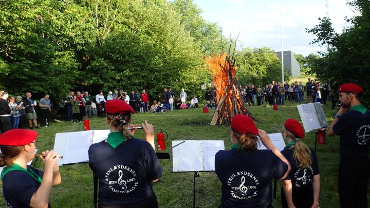 Et bål brænder, og en flok tilskuere ser til på afstand. Bag menneskemængden er der høje, grønne træer. Forrest i billedet er fem medlemmer af tambourkorpset som spiller instrumenter