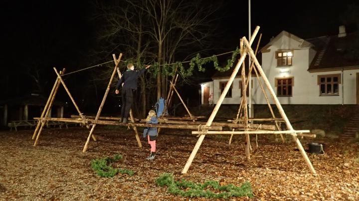 Nogle rafter er samlet til en midlertidig konstruktion, som skal bruges til at læne juletræer op ad. En spejder står på et af spærene cirka en meter over jorden og hænger en julekrans over et reb, der hænger mellem to høje punkter på spærene. En anden spejder står på jorden ved siden af ham. Himlen er mørk, og spejderpladsen er oplyst af kunstigt lys. I baggrunden kan man se et hus.