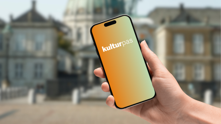 en hånd holder en mobiltelefon med "kulturpas" på skærmen, bagved er set gammel bygninger