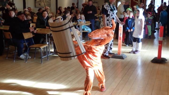 Et barn klædt ud som en orange dinosaur svinger en kæp mod en fastelavns-tønde, der holdes oppe i luften. En stor gruppe mennesker står eller sidder i baggrunden. 