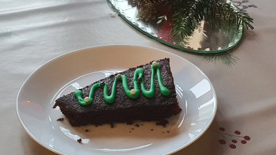 Et stykke chokoladekage formet som et juletræ ligger på en hvid tallerken. Kagen er dekoreret med grøn glasur i form af et juletræ. Ved siden af kagen står et tændt rødt stearinlys og nogle fyrrekogler på et sølvfad. Bordet er dækket med et hvidt dug med et julemønster.